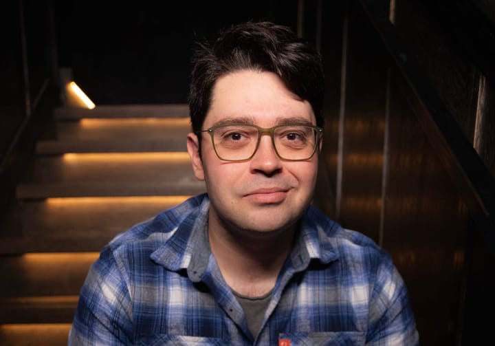 A mid 30's man sitting on a staircase wearing a blue and white flannel.